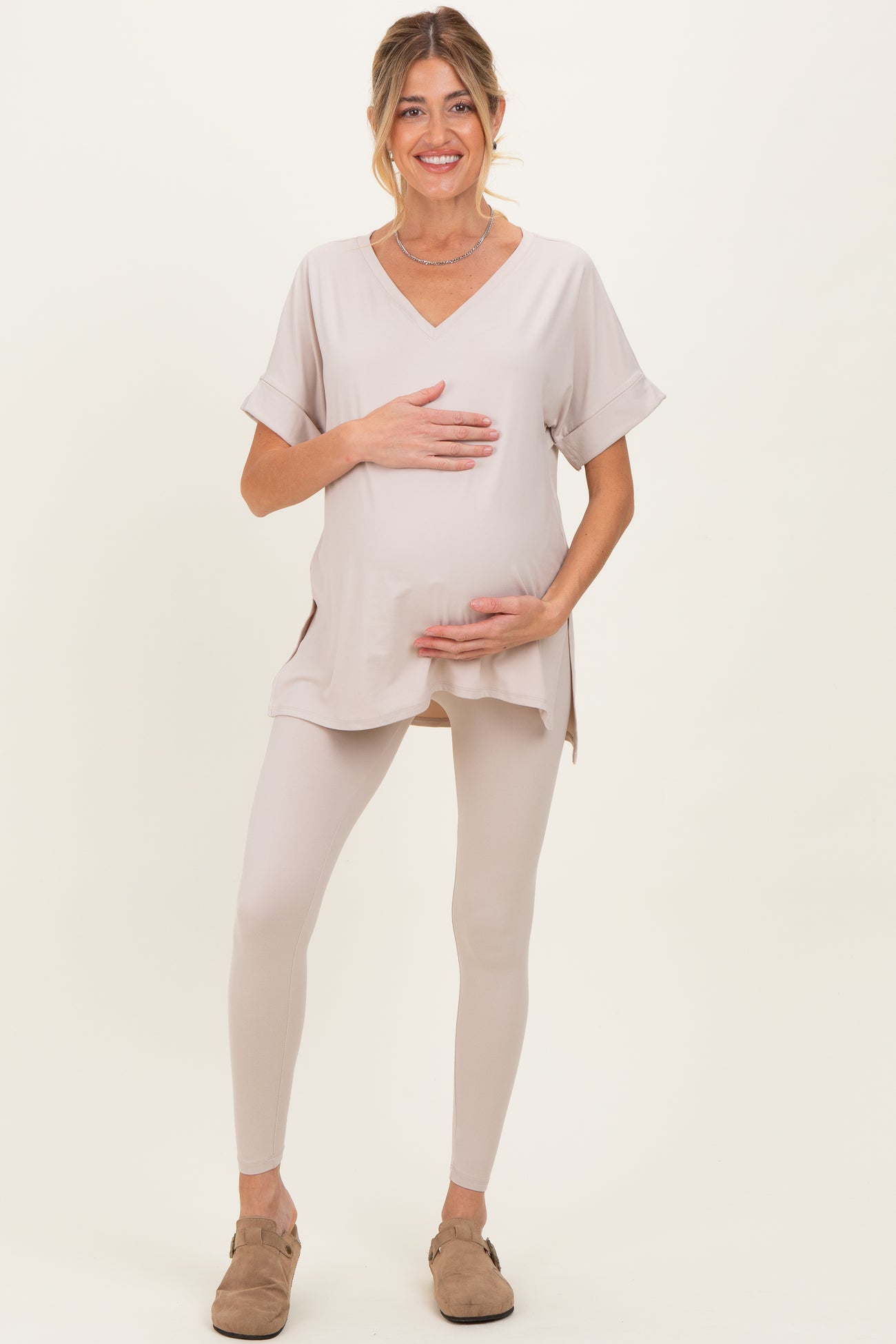 woman wearing a beige maternity top and matching leggings, holding her baby bump with a smile