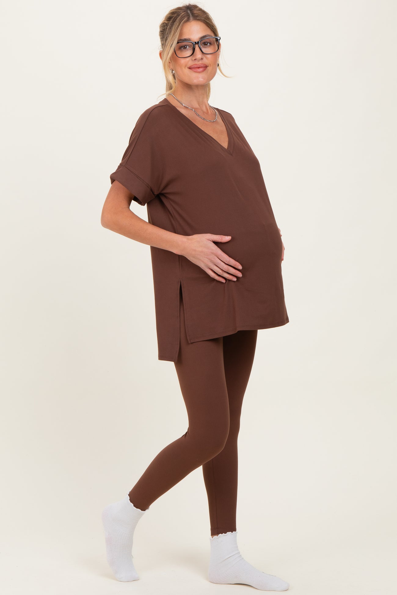 woman wearing a brown maternity top and matching leggings, gently holding her baby bump while standing sideways