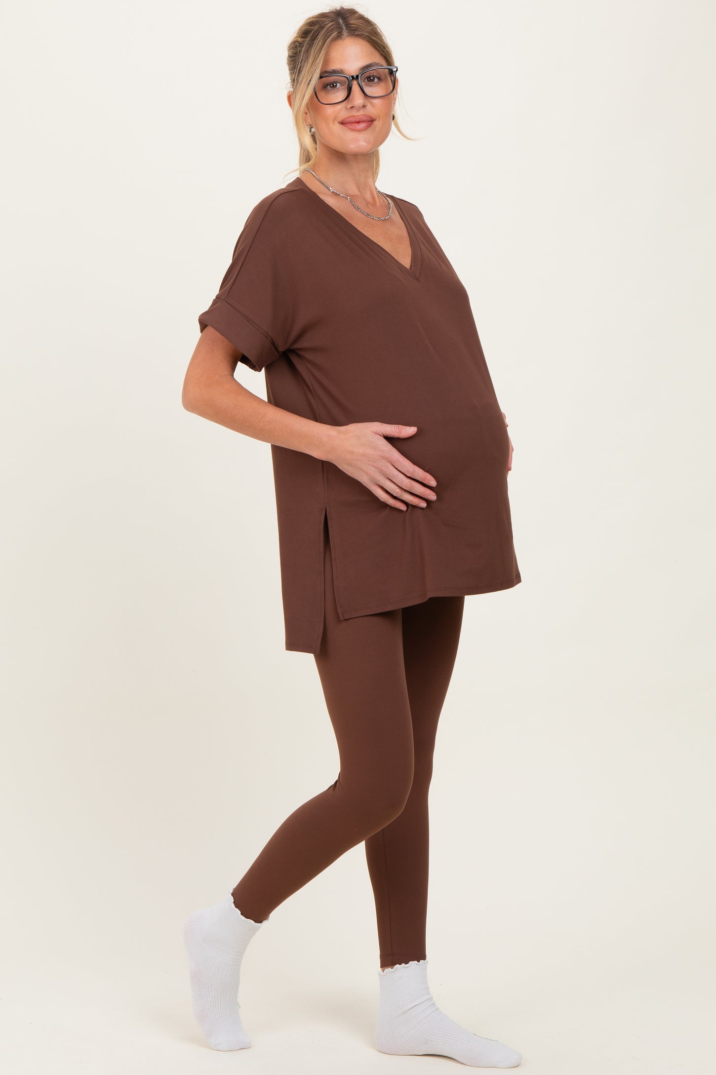 woman wearing a brown maternity top and matching leggings, gently holding her baby bump while standing sideways