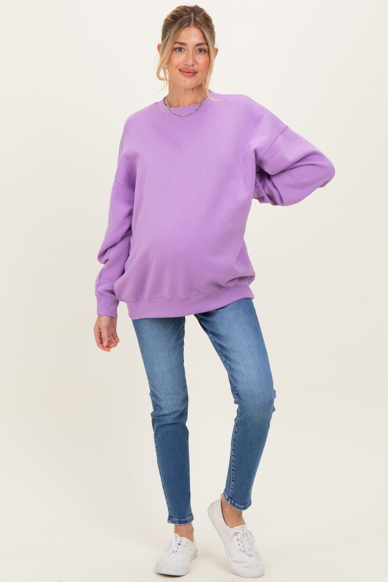 woman wearing a light purple oversized maternity sweatshirt and blue skinny jeans, showing her baby bump