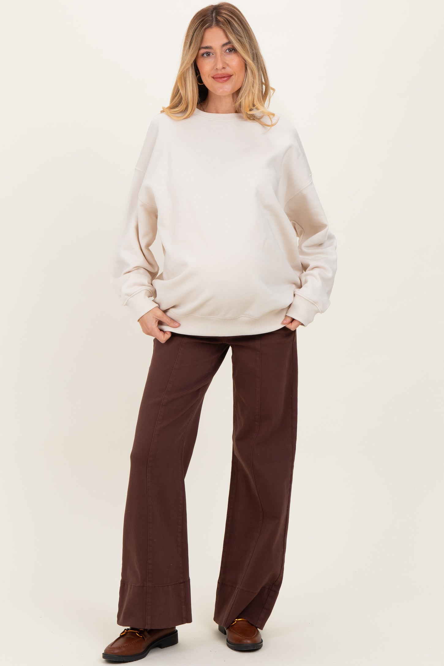woman wearing a beige maternity sweatshirt and brown wide-leg pants, showcasing her baby bump