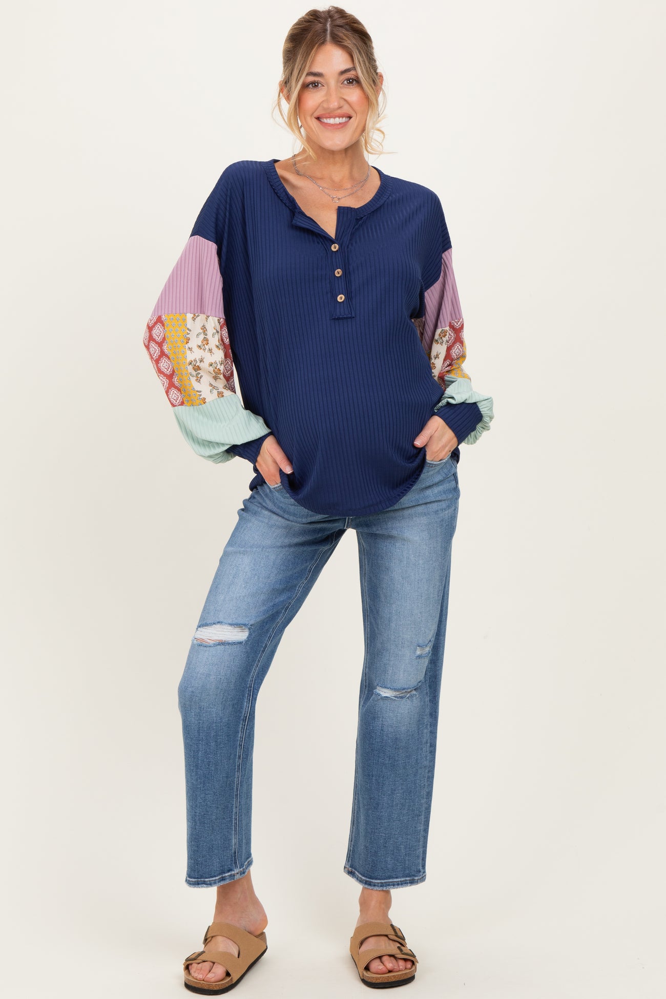 woman wearing a navy blue maternity top with patterned long sleeves and distressed jeans, smiling and standing confidently