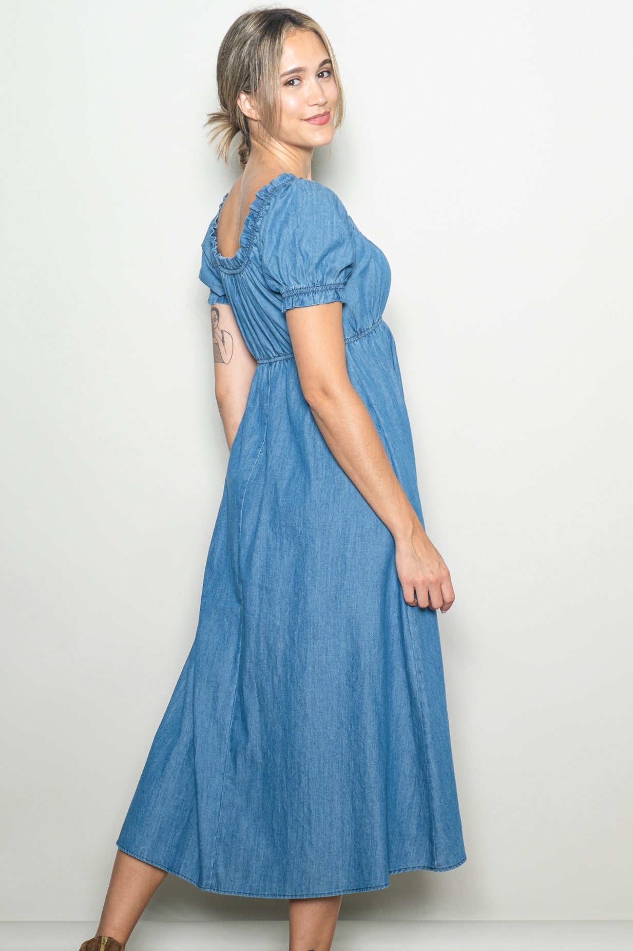 woman wearing a blue denim maxi dress with puff sleeves and a gathered back detail, smiling over her shoulder