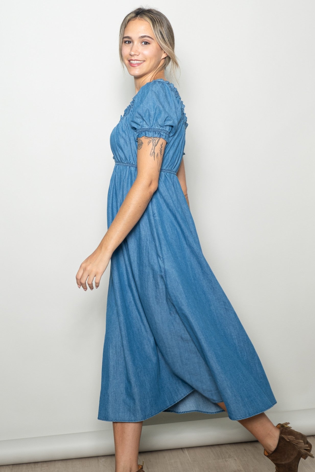 woman wearing a blue denim maxi dress with short puff sleeves and a ruched bodice, smiling while standing sideways