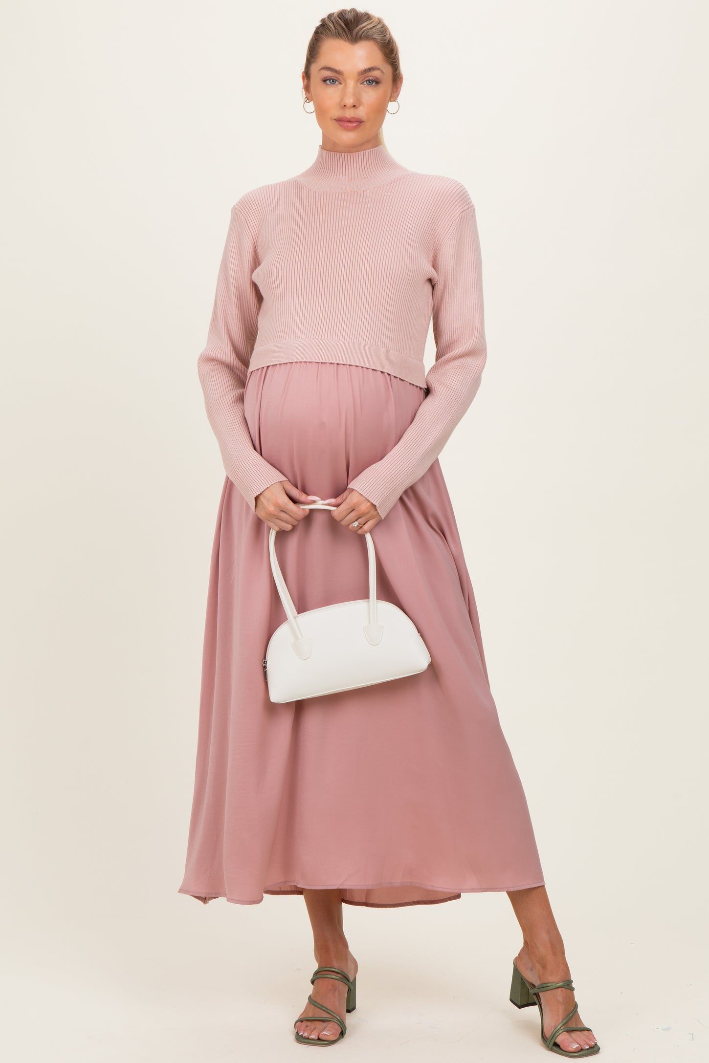 woman wearing a light pink ribbed maternity sweater and a flowy mauve maxi skirt, holding a white handbag and showing her baby bump
