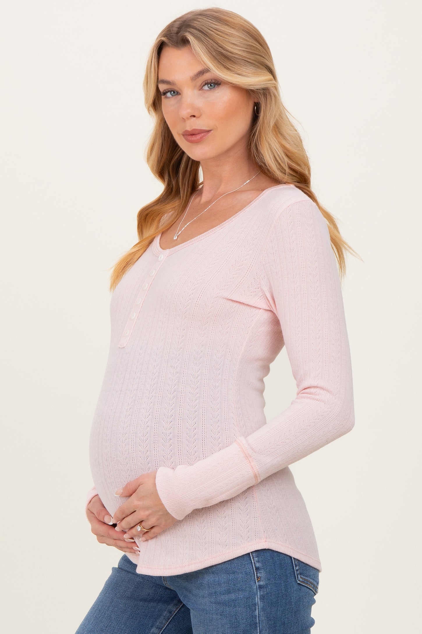 woman wearing a light pink long sleeve maternity top with a henley neckline and holding her baby bump
