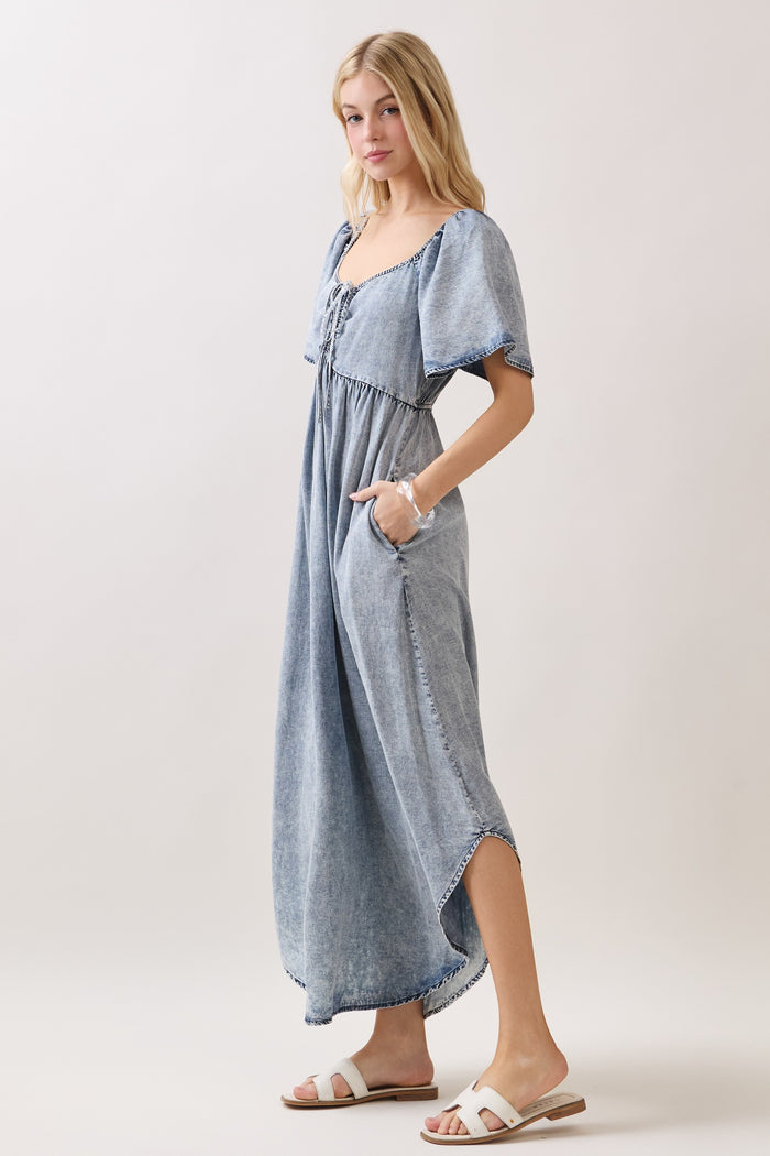 woman wearing a light blue denim maxi dress with short sleeves, pockets, and lace-up front details