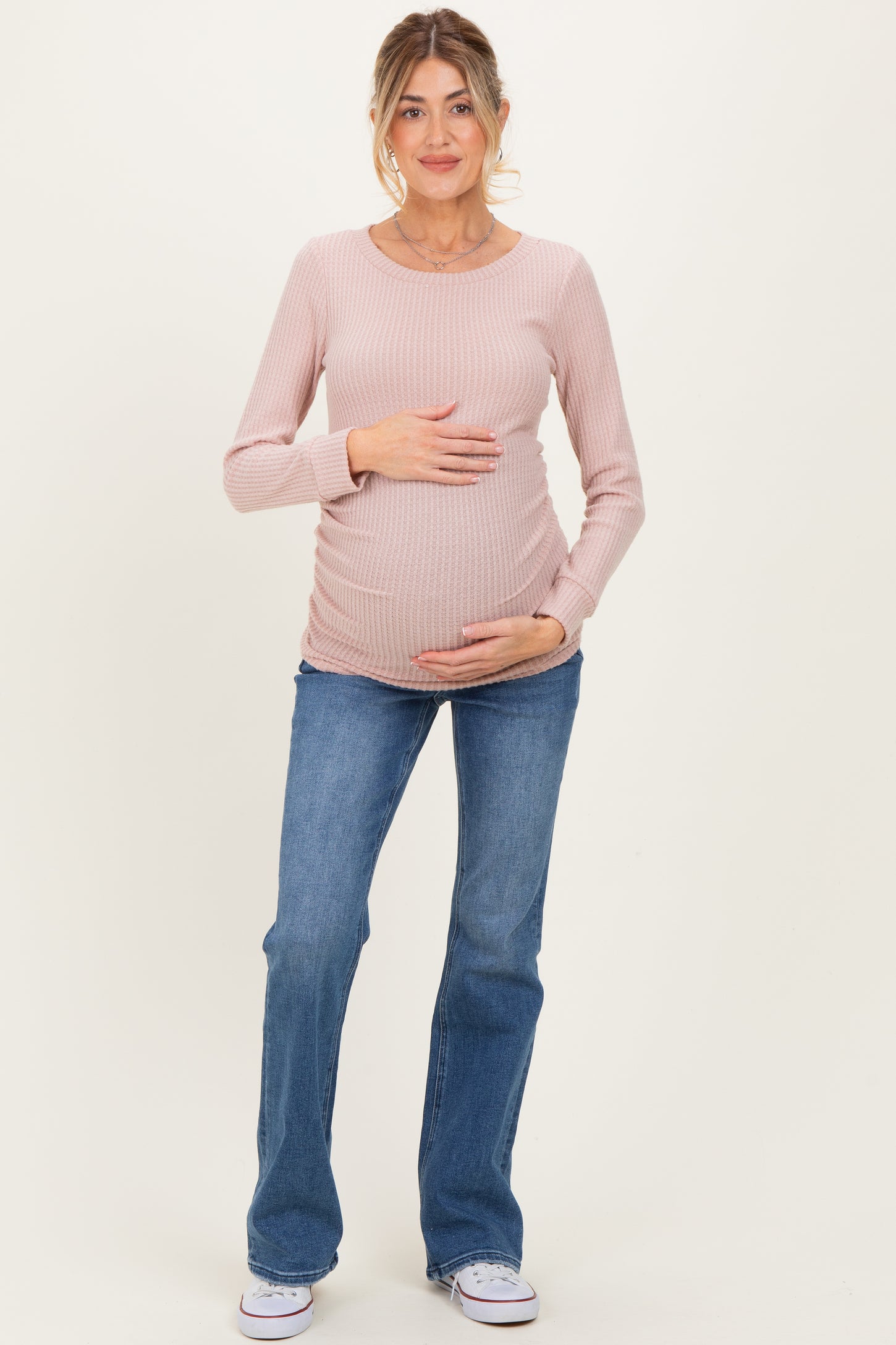 woman wearing a light pink long sleeve ribbed maternity top and blue flared jeans, gently holding her baby bump