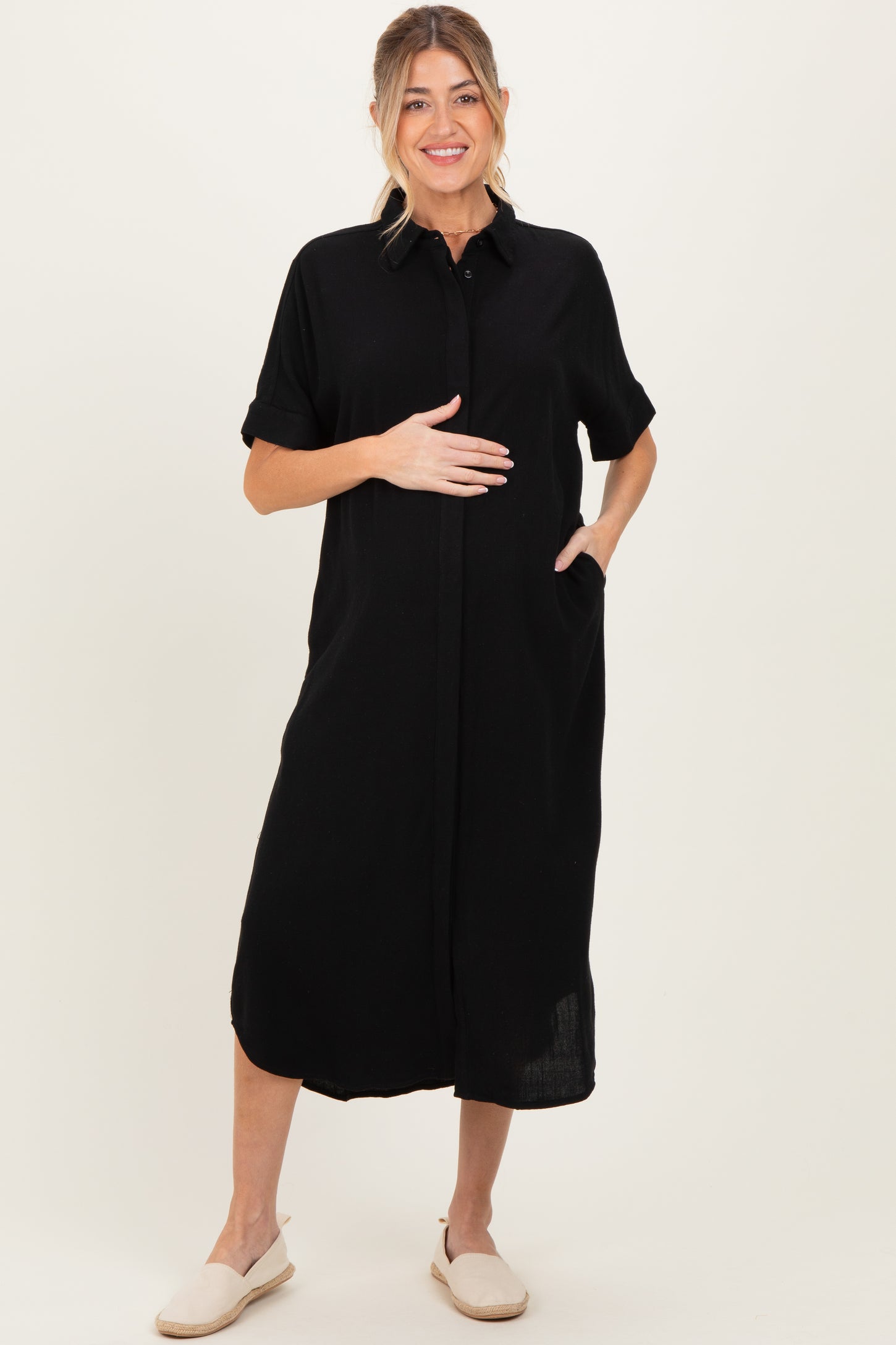 woman wearing a black short sleeve maternity dress with a button-up collar and pockets, holding her baby bump
