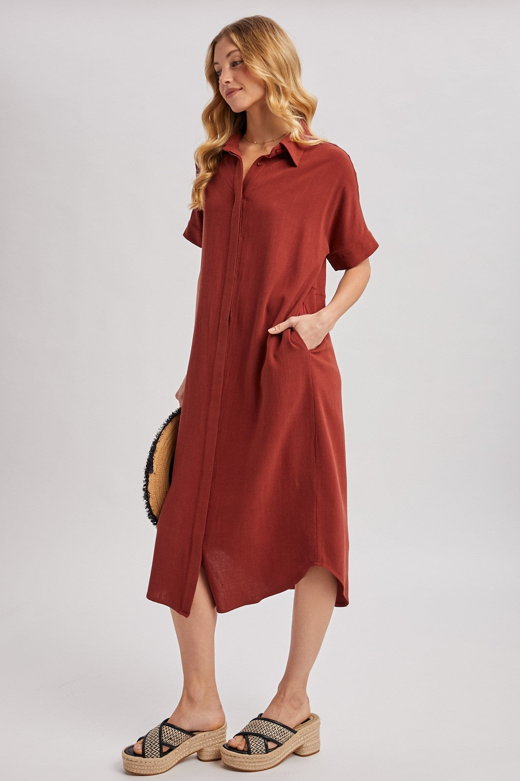 woman wearing a rust short sleeve button-up dress with pockets and a high-low hem, styled with sandals and a round bag