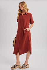 woman wearing a rust short sleeve button-up dress with pockets and a high-low hem, styled with sandals and a round bag