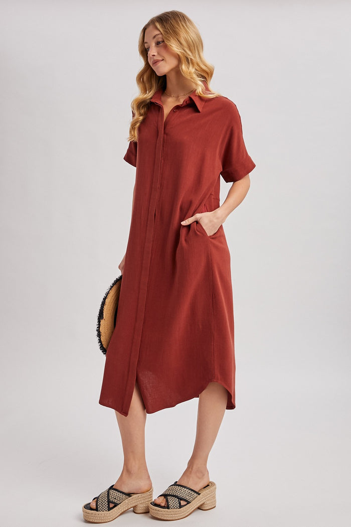 woman wearing a rust short sleeve button-up dress with pockets and a high-low hem, styled with sandals and a round bag