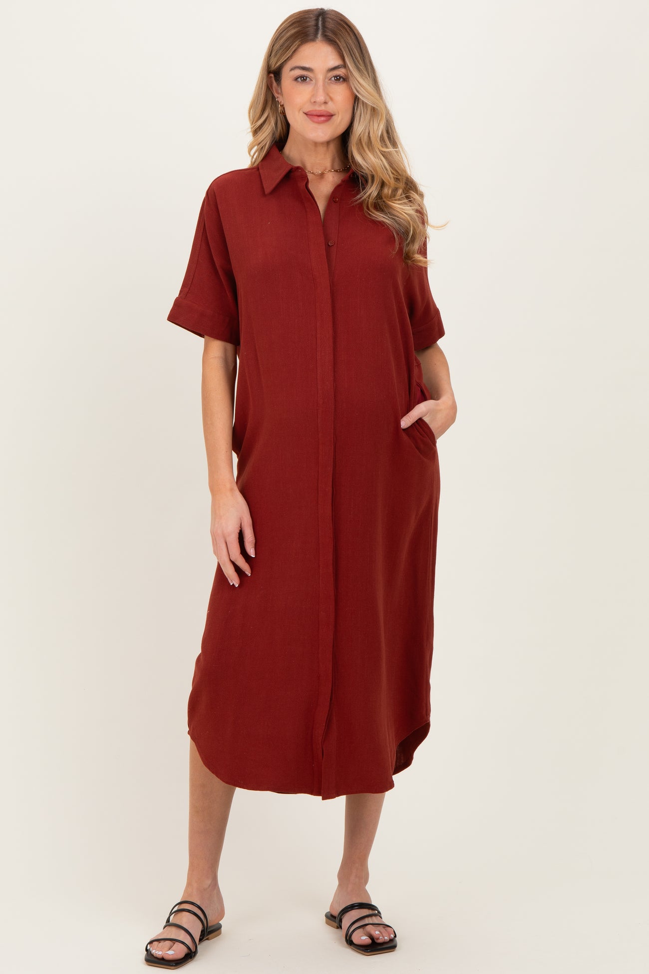 woman wearing a rust-colored maternity shirt dress with short sleeves and button front, posing with hands in pockets
