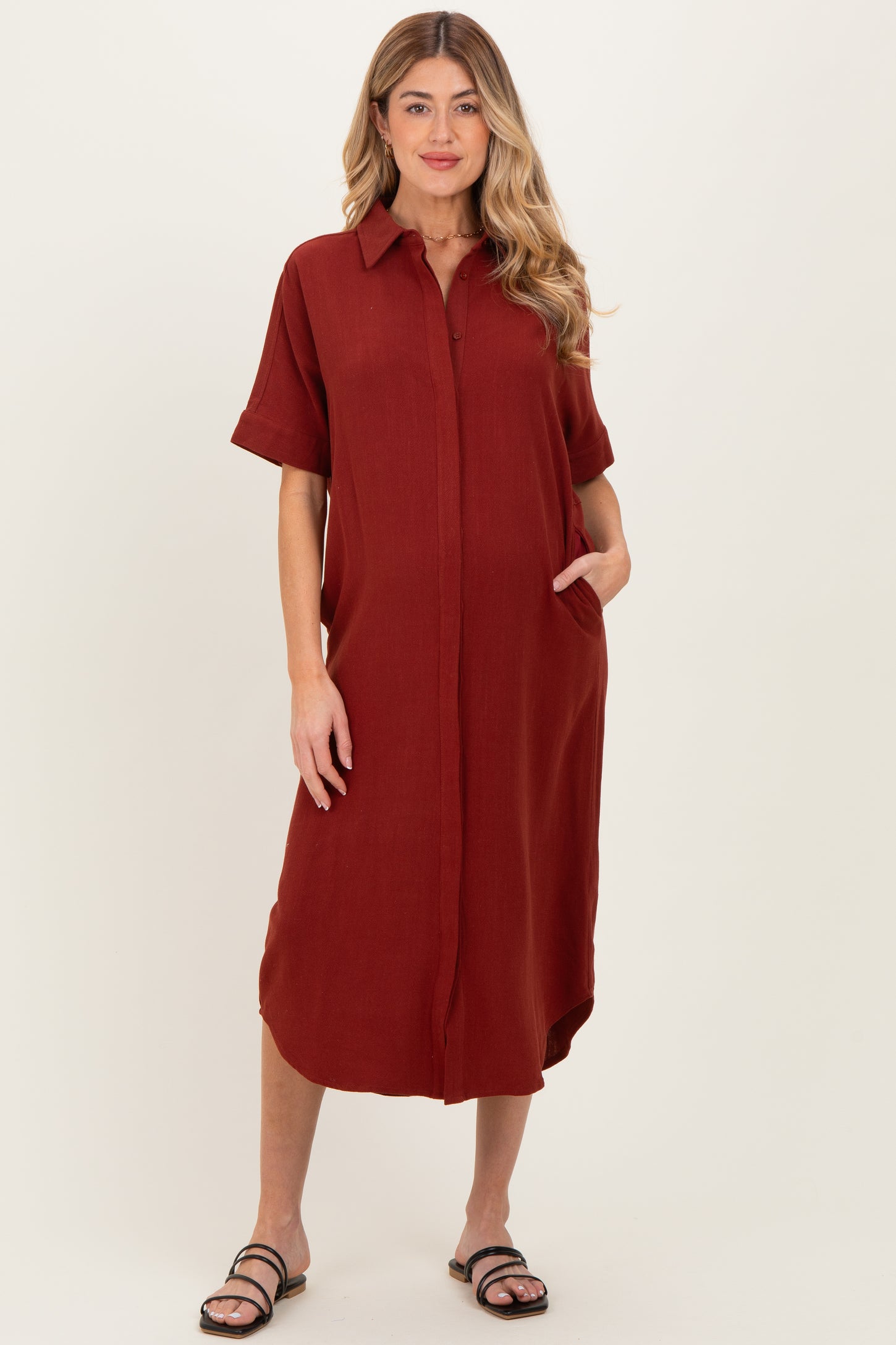 woman wearing a rust-colored maternity shirt dress with short sleeves and button front, posing with hands in pockets