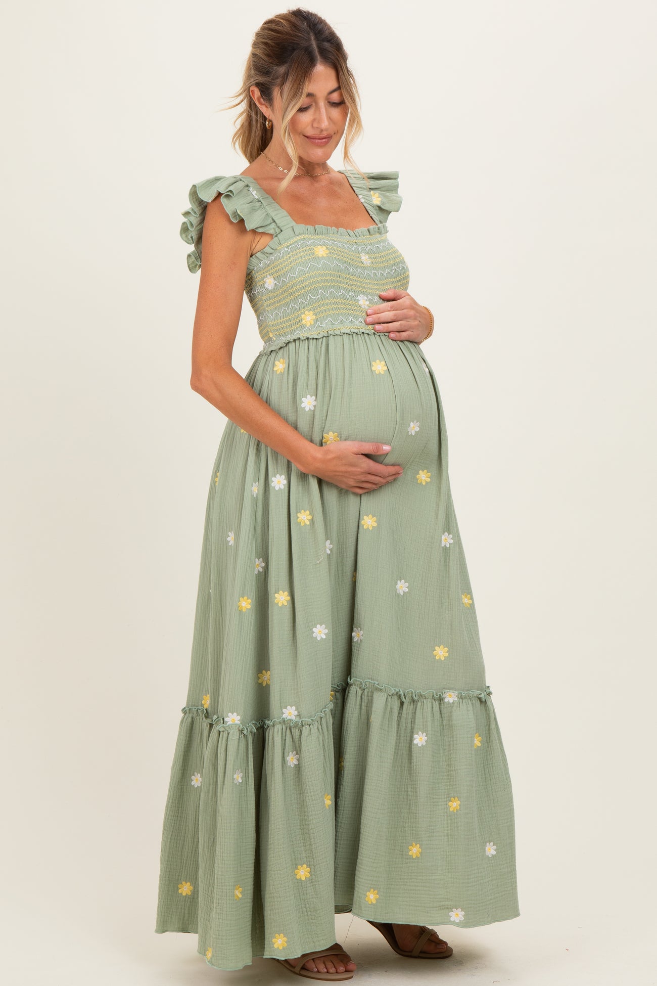 woman wearing a light green maternity maxi dress with floral embroidery and ruffled straps, cradling her baby bump