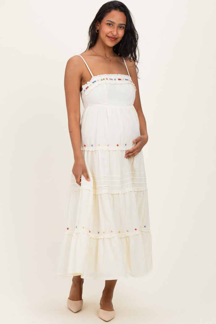 woman wearing a white maternity maxi dress with colorful embroidery and a baby bump, smiling and posing with one hand on her belly