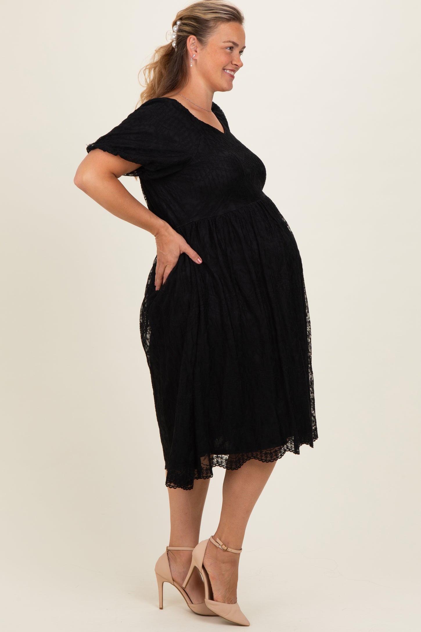 woman wearing a black lace maternity dress and showing her baby bump, paired with beige high-heeled shoes