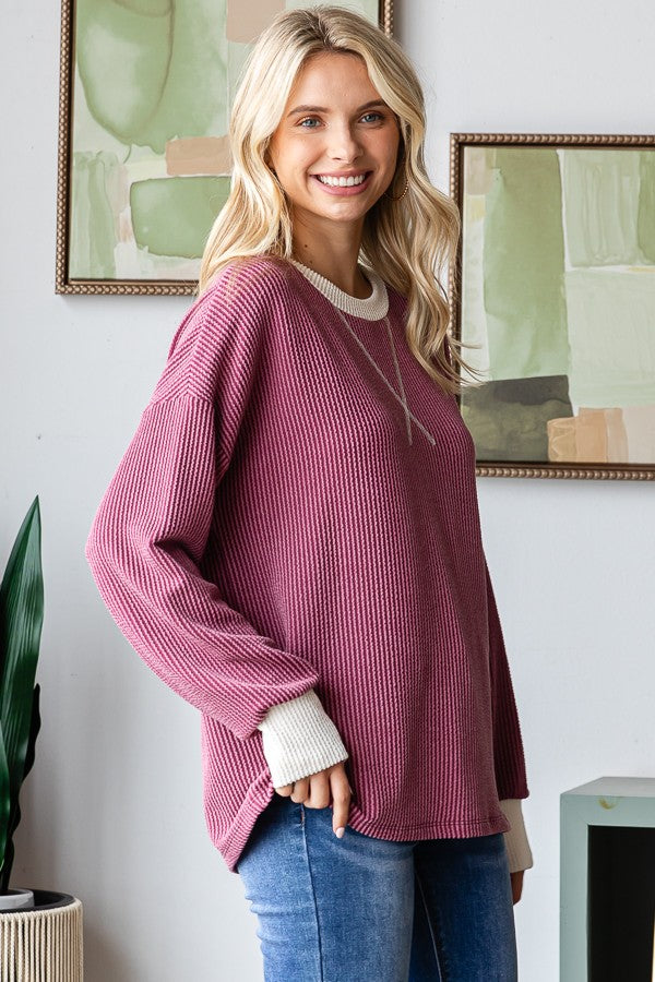 woman wearing a burgundy ribbed oversized sweater with cream cuffs and a relaxed fit, smiling against a decorative background
