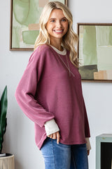 woman wearing a burgundy ribbed oversized sweater with cream cuffs and a relaxed fit, smiling against a decorative background