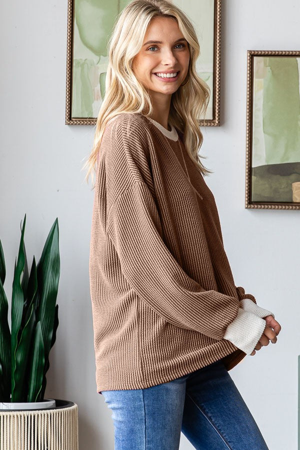 woman wearing a brown oversized ribbed sweater with cream cuff details and blue jeans, standing in a bright room