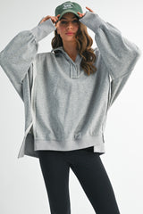 woman wearing a gray oversized hoodie with a half-zip and wide sleeves, paired with black leggings and a green baseball cap