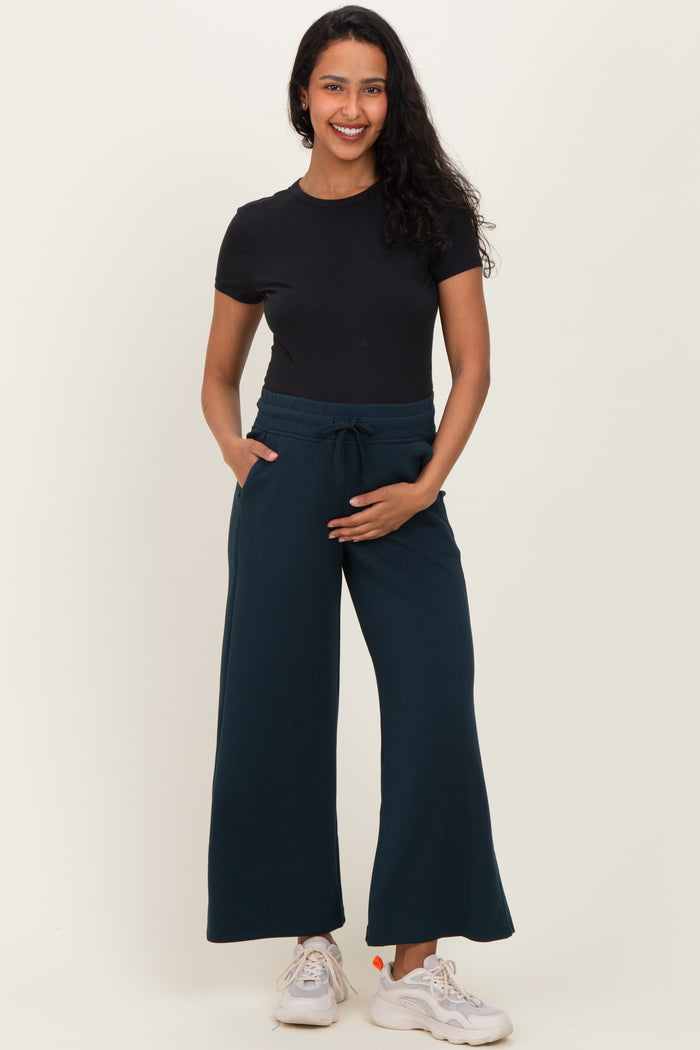 woman wearing a black short sleeve top and dark teal maternity wide-leg pants, holding her baby bump with one hand