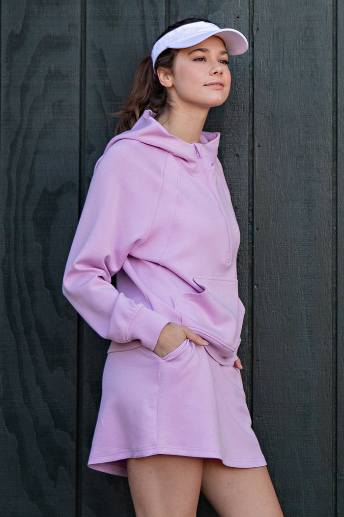 woman wearing a pastel pink sweatshirt dress with a hood and pockets, paired with a white visor cap