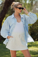 woman wearing a light blue oversized button-up shirt and gray maternity mini skirt, holding sunglasses outdoors