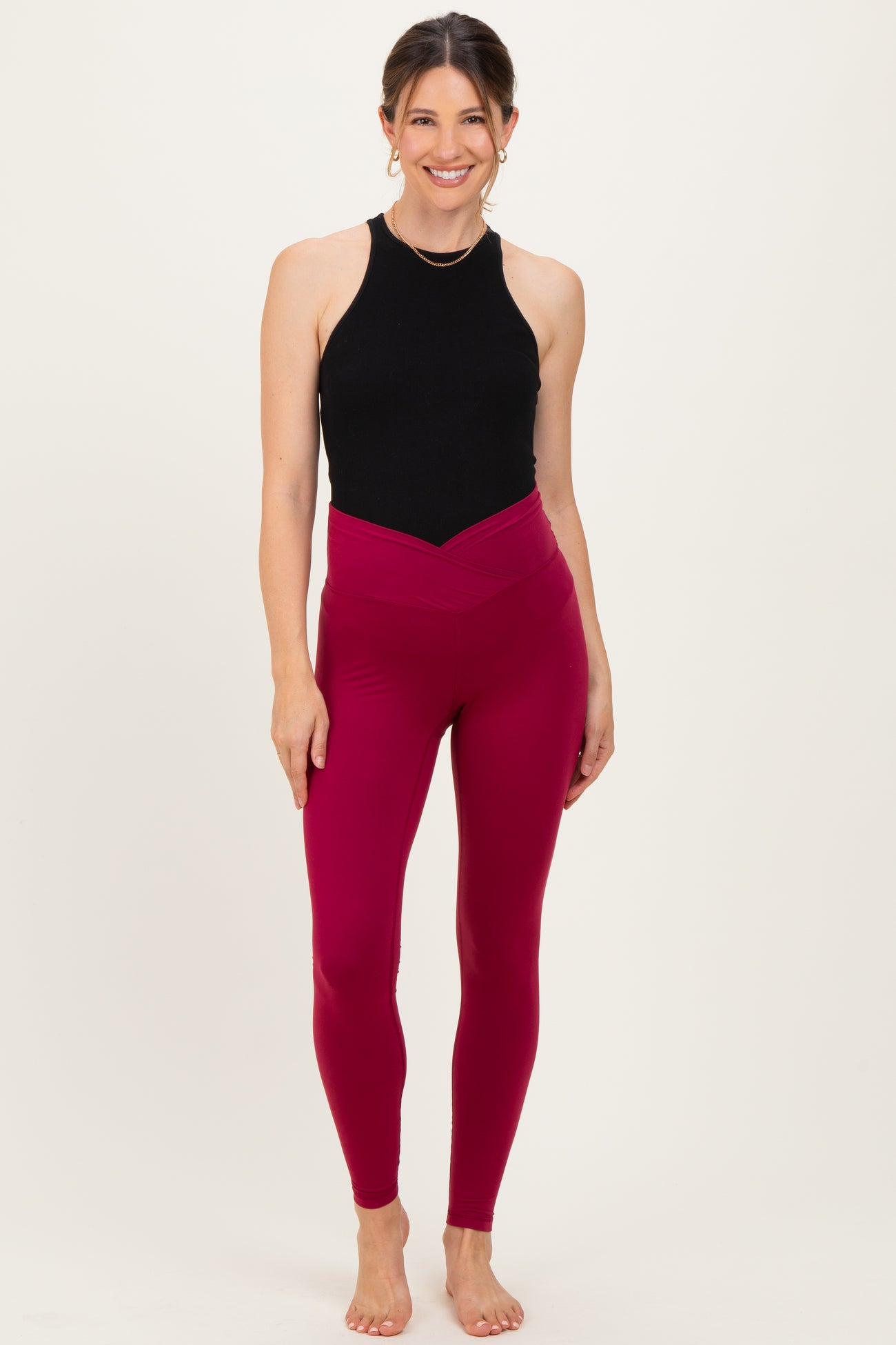 woman wearing a black sleeveless maternity top and burgundy high-waisted maternity leggings, standing barefoot
