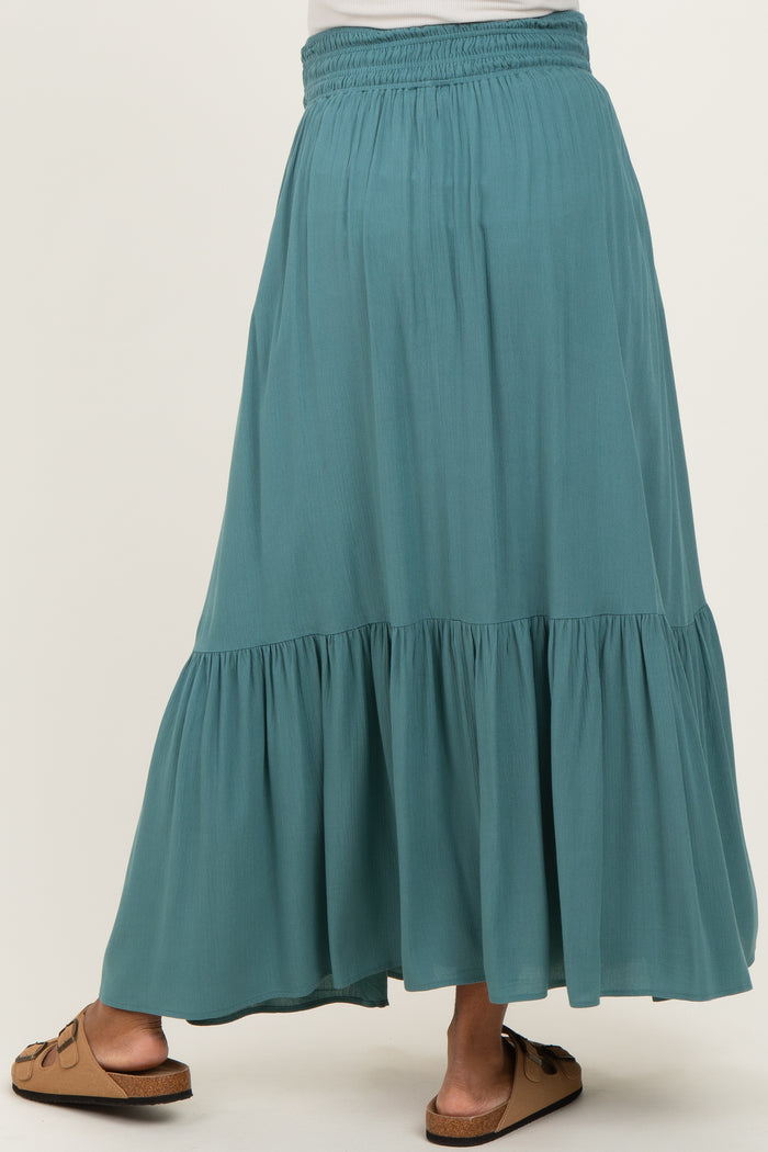 woman wearing a teal maternity maxi skirt with a tiered design and elastic waistband, standing with her back to the camera