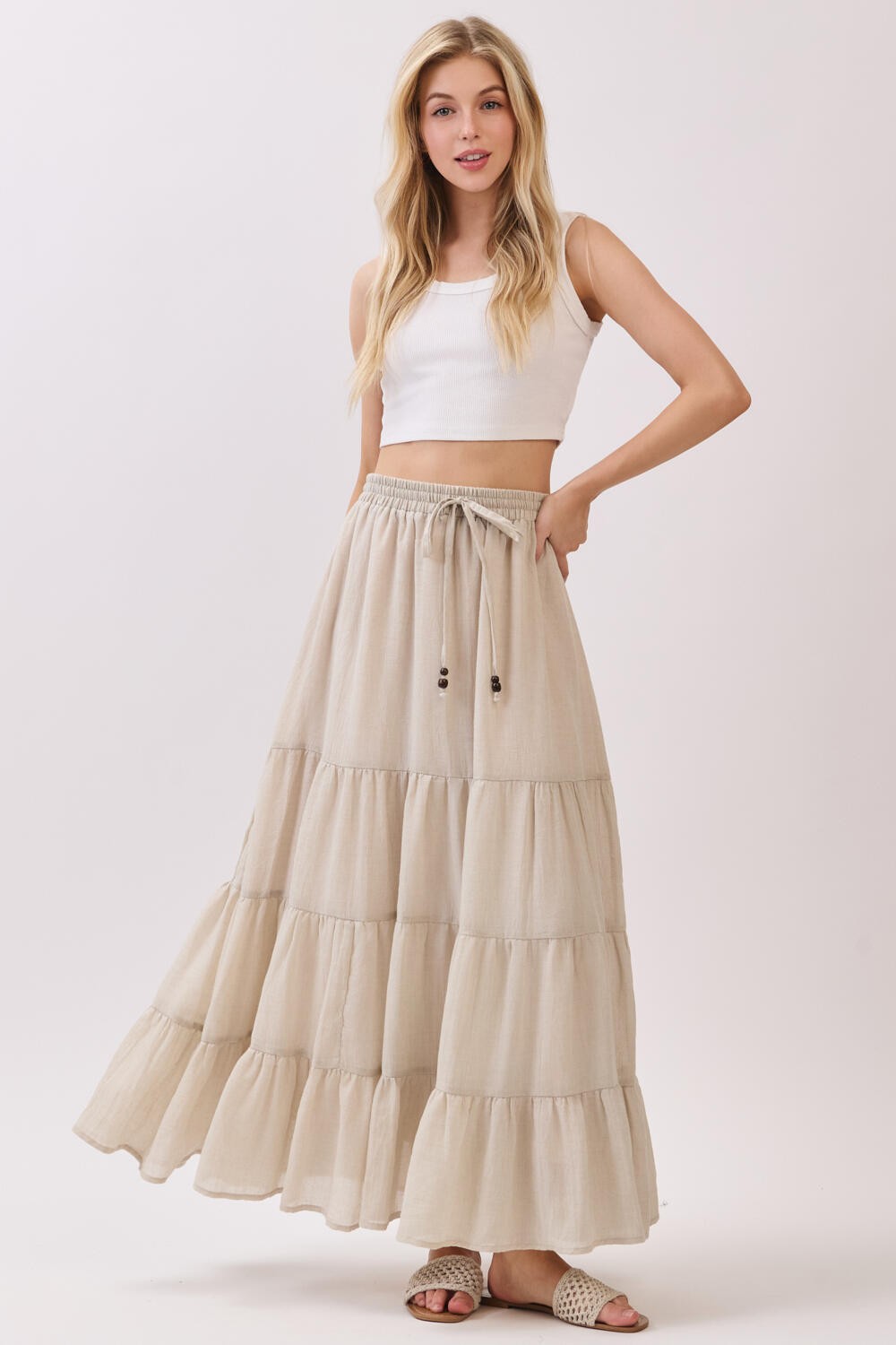 woman wearing a cream tiered maxi skirt and a white cropped tank top, smiling while holding the skirt's waistband