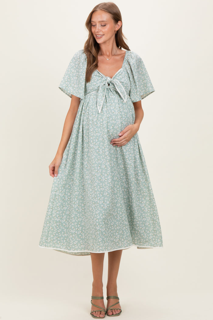 woman wearing a light green floral print maternity dress with a tie front, holding her baby bump