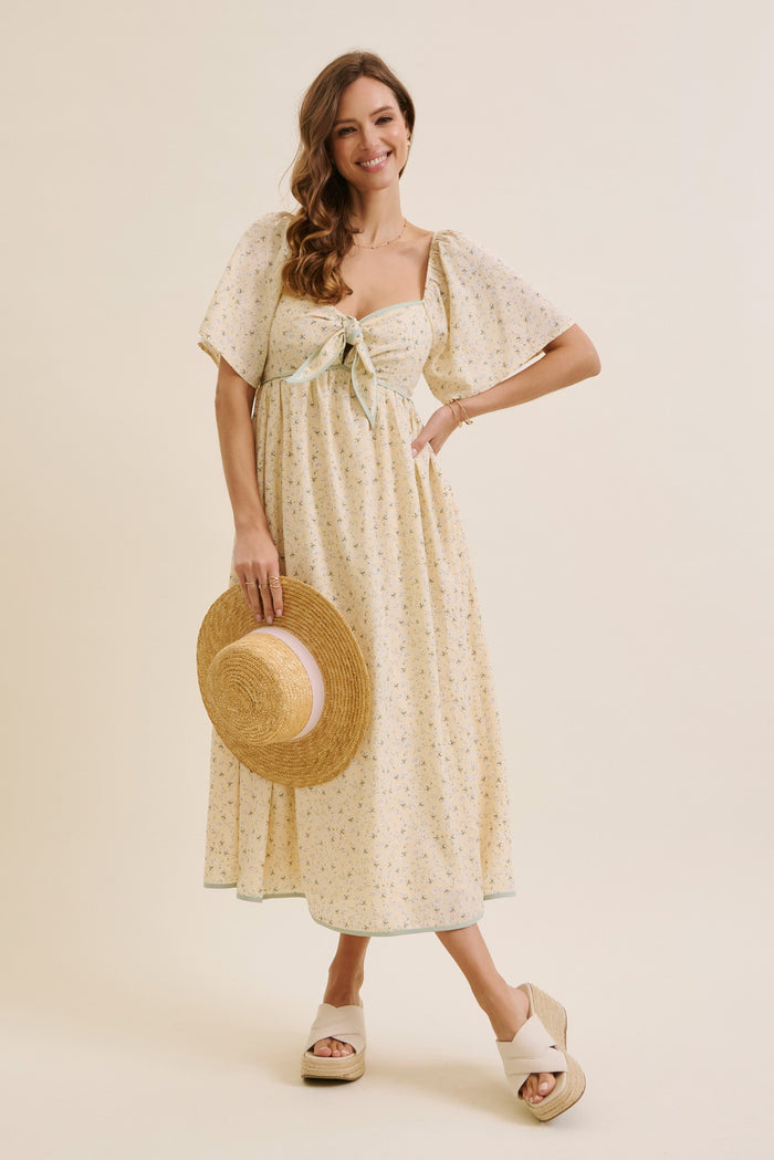 woman wearing a yellow floral midi dress with puff sleeves and a tie front, holding a straw hat and wearing wedge sandals