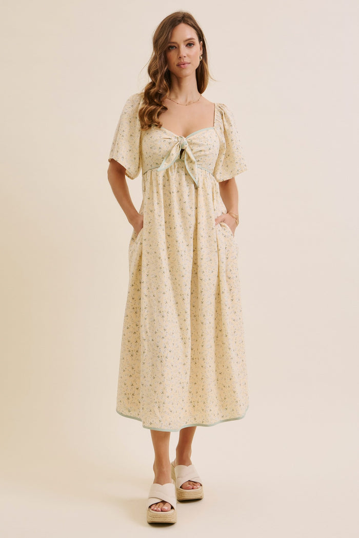 woman wearing a light yellow floral midi dress with short puff sleeves and a tie front, standing with hands in pockets