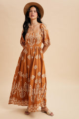 woman wearing a rust-colored maxi dress with white floral patterns and puff sleeves, paired with a straw hat and sandals