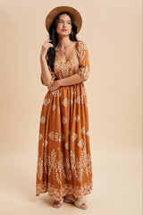 woman wearing a brown maxi dress with white floral patterns and a straw hat, posing with hand on her hip