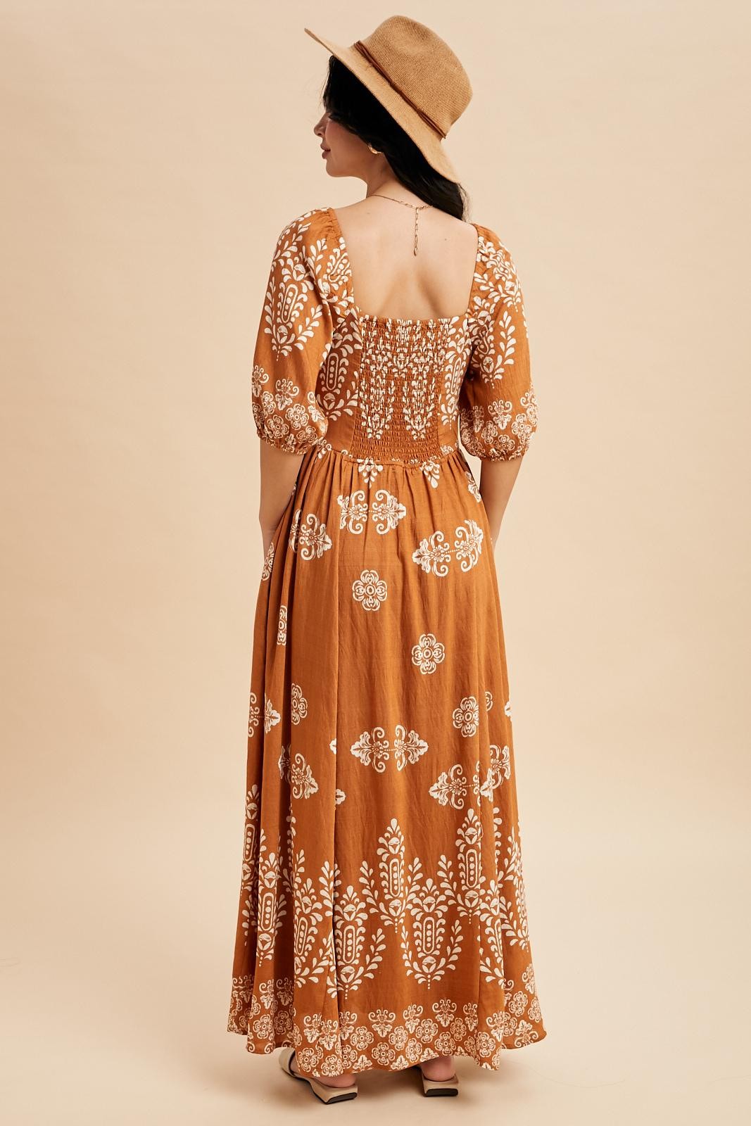 woman wearing a rust-colored maxi dress with white floral patterns and puff sleeves, standing with back to camera and wearing a hat