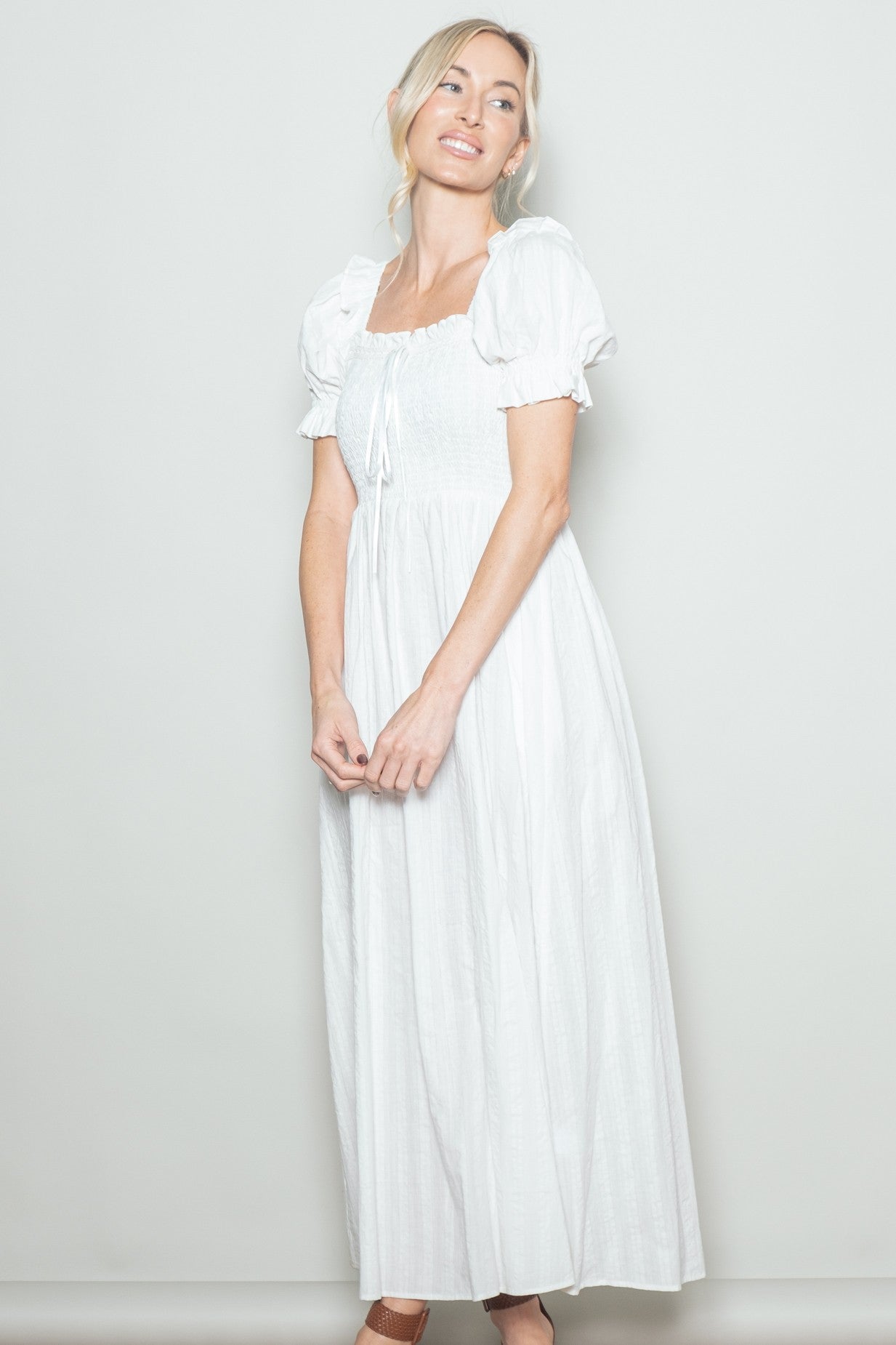 woman wearing a white maxi dress with puffy sleeves and a smocked bodice, smiling against a neutral background