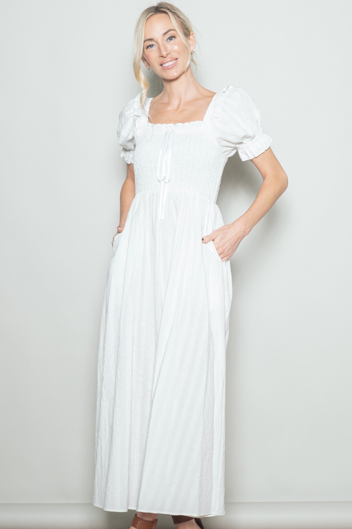woman wearing a white maxi dress with puff sleeves and a smocked bodice, featuring pockets and a tie at the front