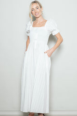 woman wearing a white maxi dress with puff sleeves and a smocked bodice, featuring pockets and a tie at the front