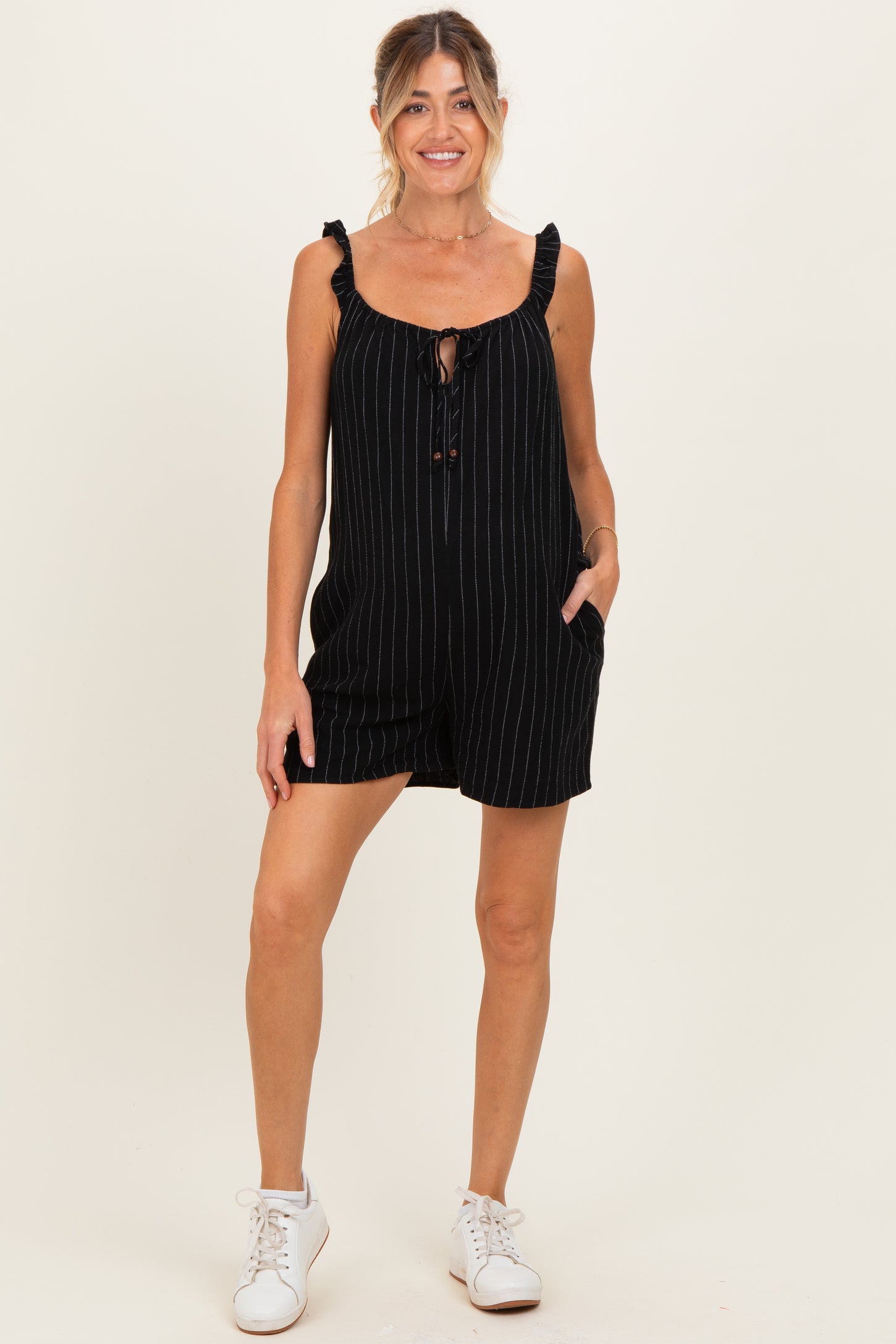 woman wearing a black striped maternity romper with ruffled straps and pockets, styled with white sneakers