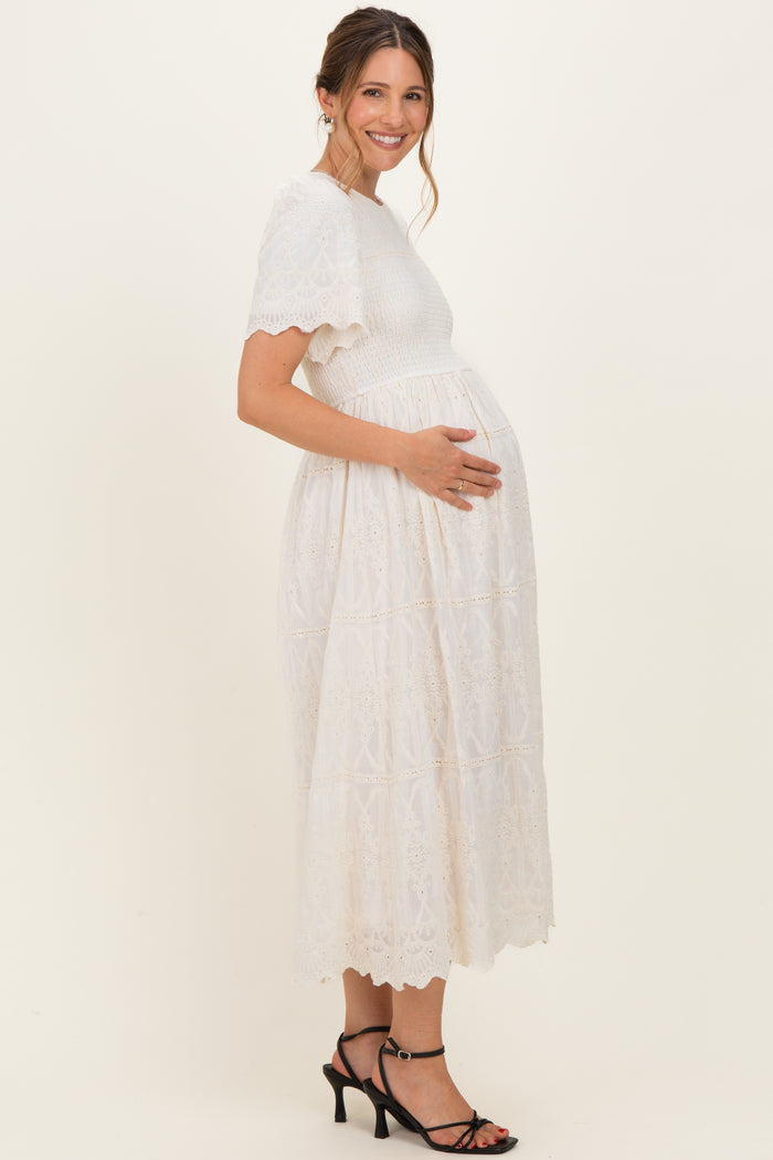 woman wearing a white lace maternity dress, smiling and holding her baby bump