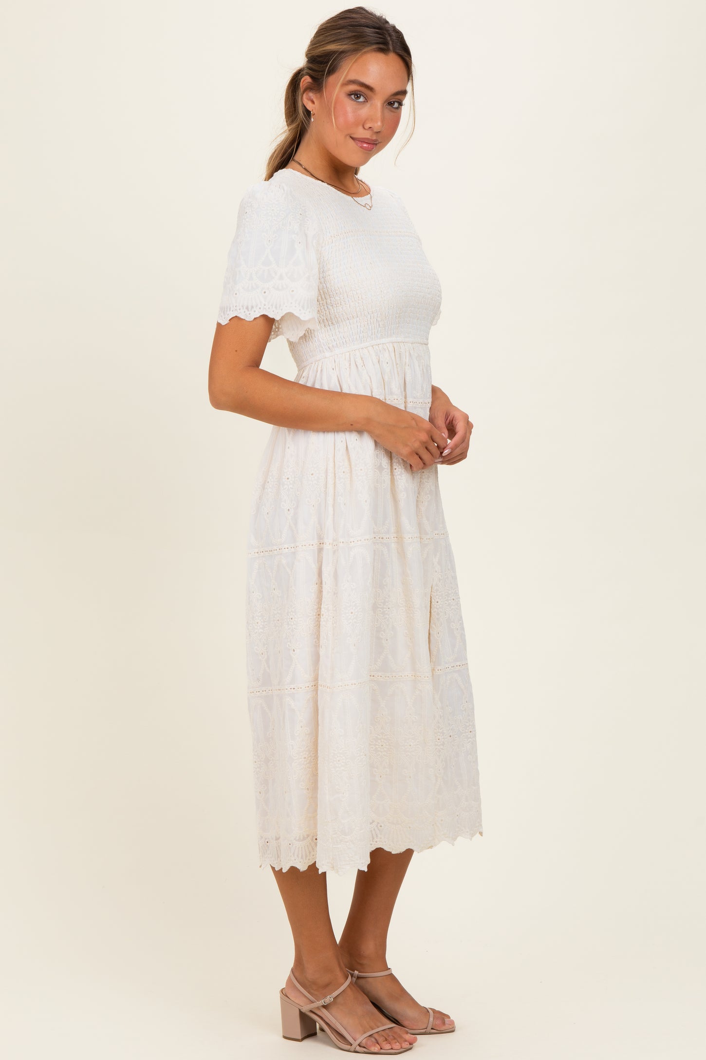 woman wearing a white embroidered midi dress with short sleeves and a cinched waist, paired with strappy sandals