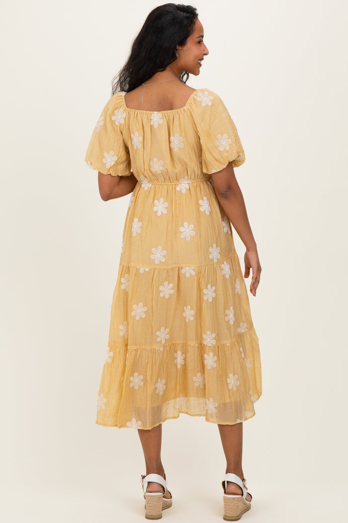 woman wearing a flowy yellow maternity dress with white floral embroidery and puff sleeves, showing off her baby bump from the back