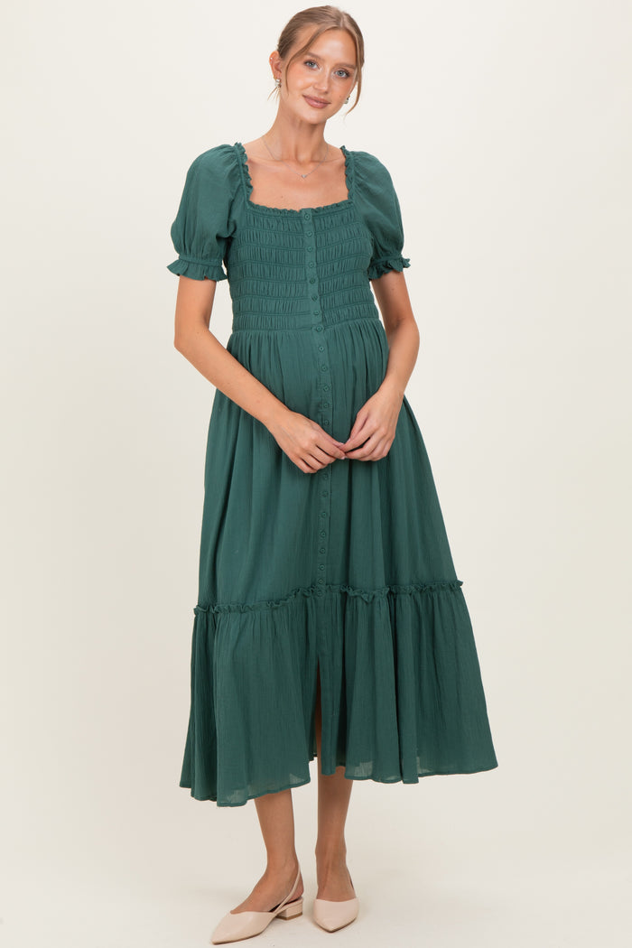 woman wearing a green maternity midi dress with puff sleeves and a smocked bodice, gently holding her baby bump