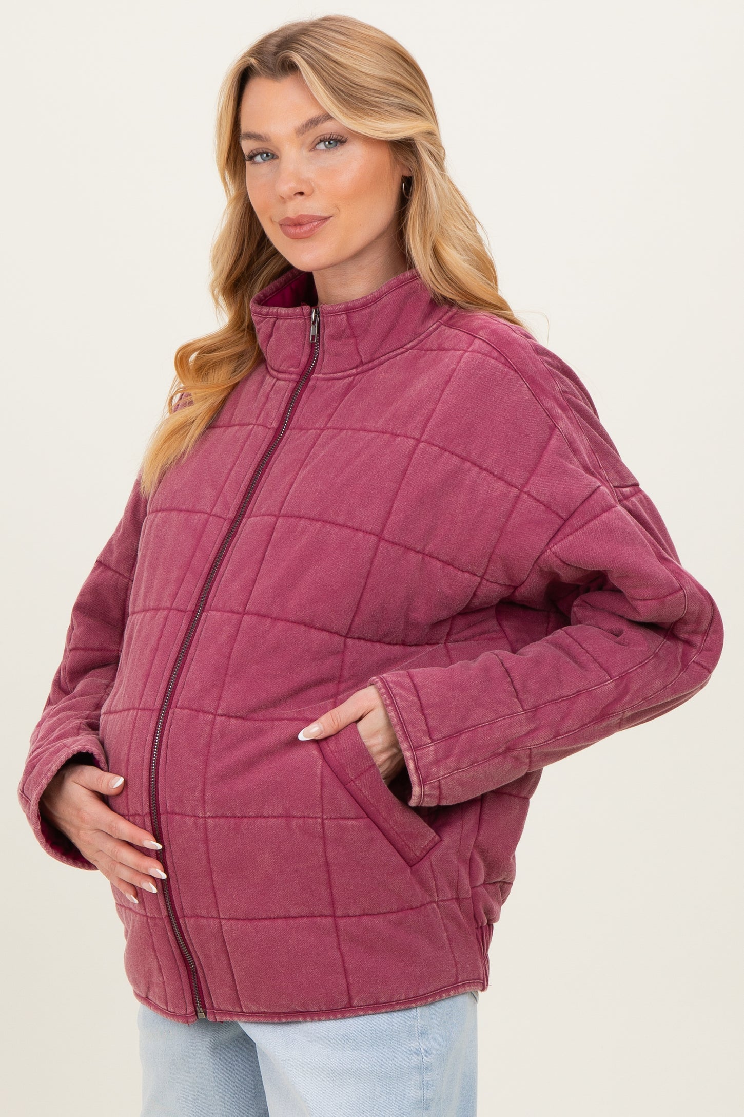 woman wearing a burgundy quilted maternity jacket with a standing collar and side pockets, gently cradling her baby bump