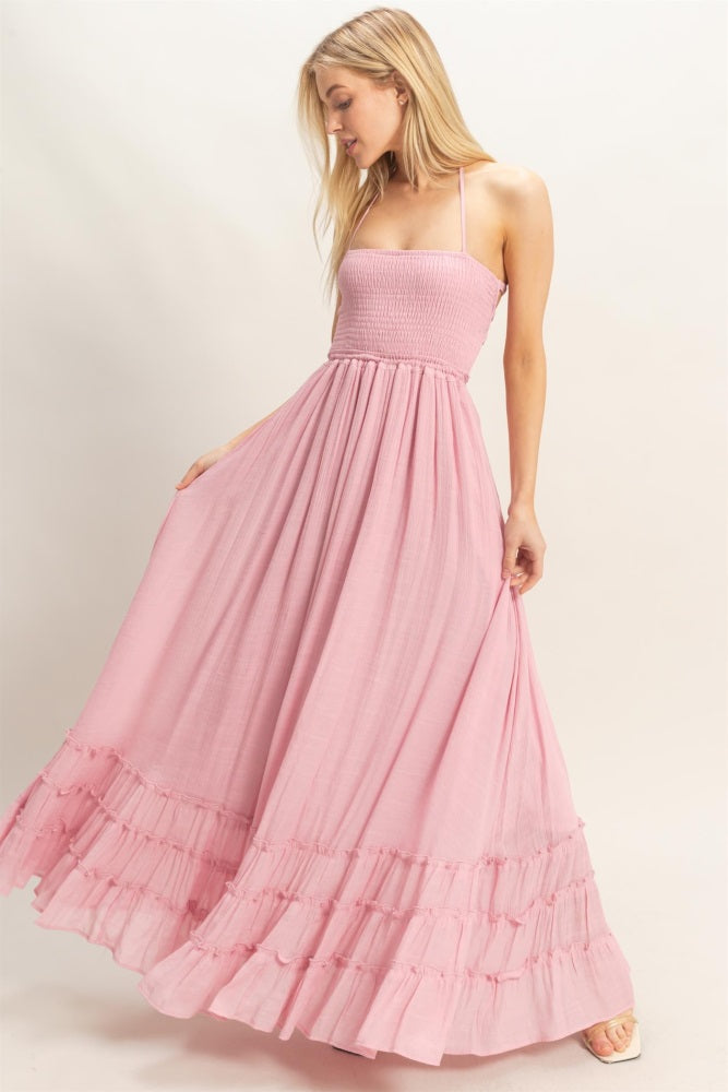 woman wearing a pink maxi dress with a smocked bodice and ruffled hem, standing and holding the skirt with both hands
