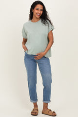 woman wearing a light green maternity top and blue rolled-up jeans, holding her baby bump
