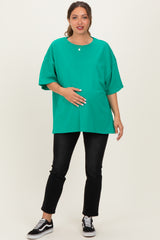 woman wearing a bright green oversized maternity top and black jeans, gently holding her baby bump