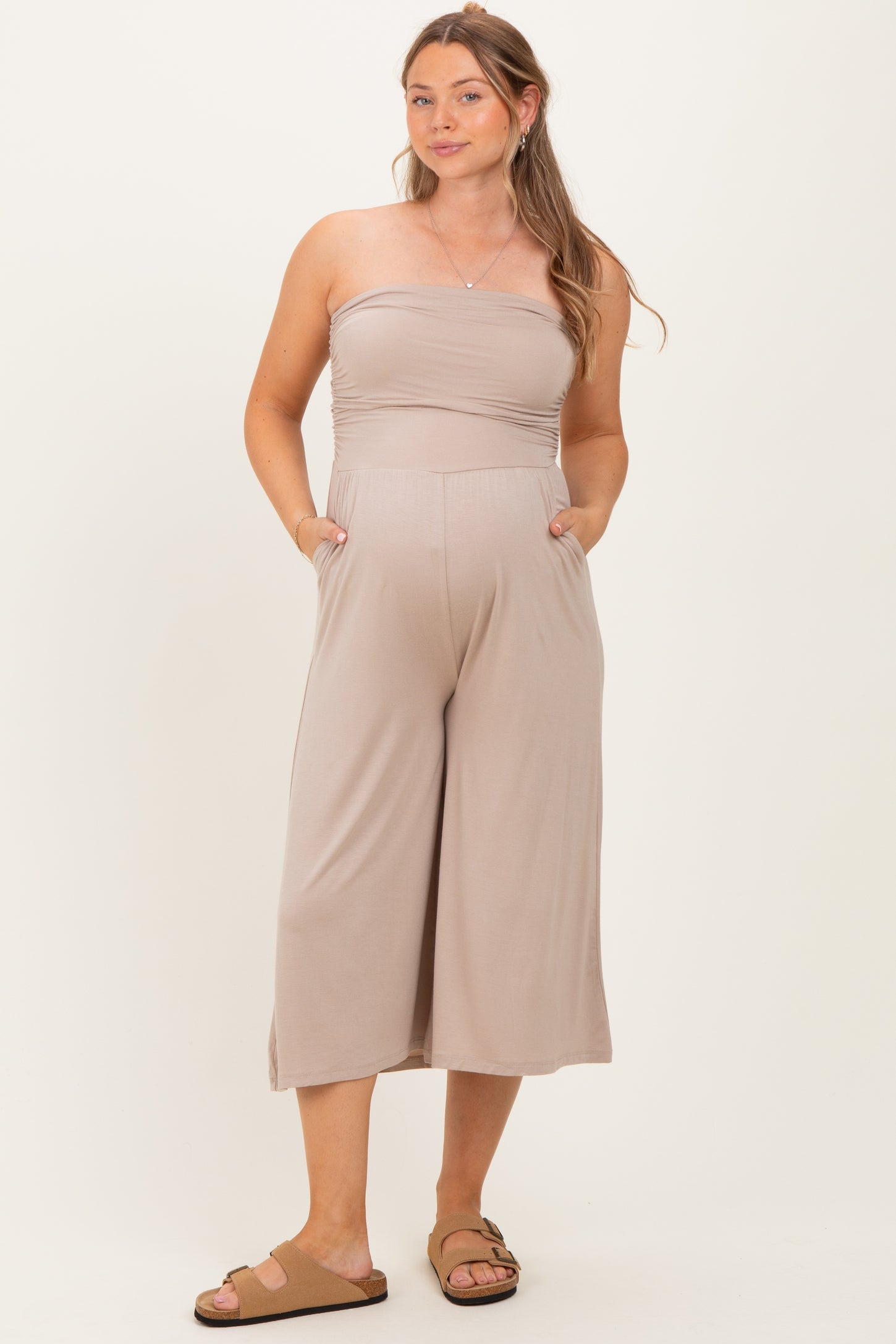 woman wearing a taupe strapless maternity jumpsuit with pockets and a flowing silhouette, showing her baby bump
